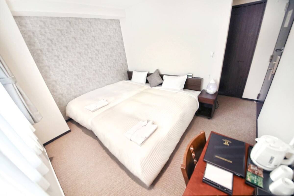 A hotel room with a large bed, a bedside table with a lamp, a small table with a book and a kettle, and a door.