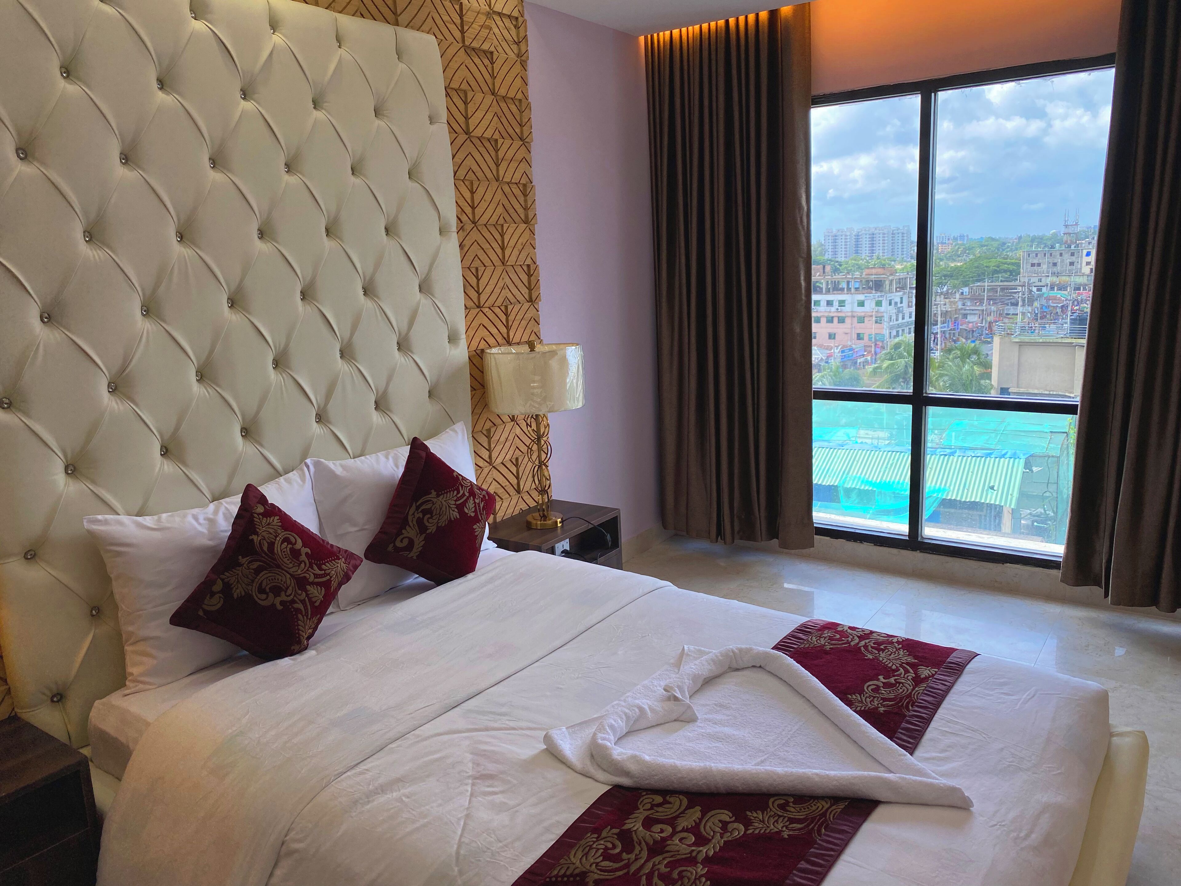 Ein Hotelzimmer mit einem gebogenem Kopfteil, einem großen Bett mit weißer und roter Bettwäsche, einem Nachttisch, einer Lampe und Blick auf die Stadt durch ein großes Fenster.