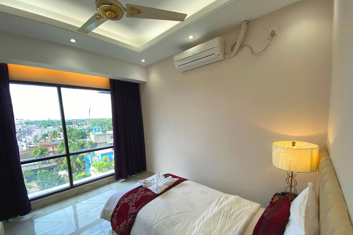 A hotel room with a large window, a bed with a red and white bedspread, a bedside lamp, and an air conditioning unit on the wall.