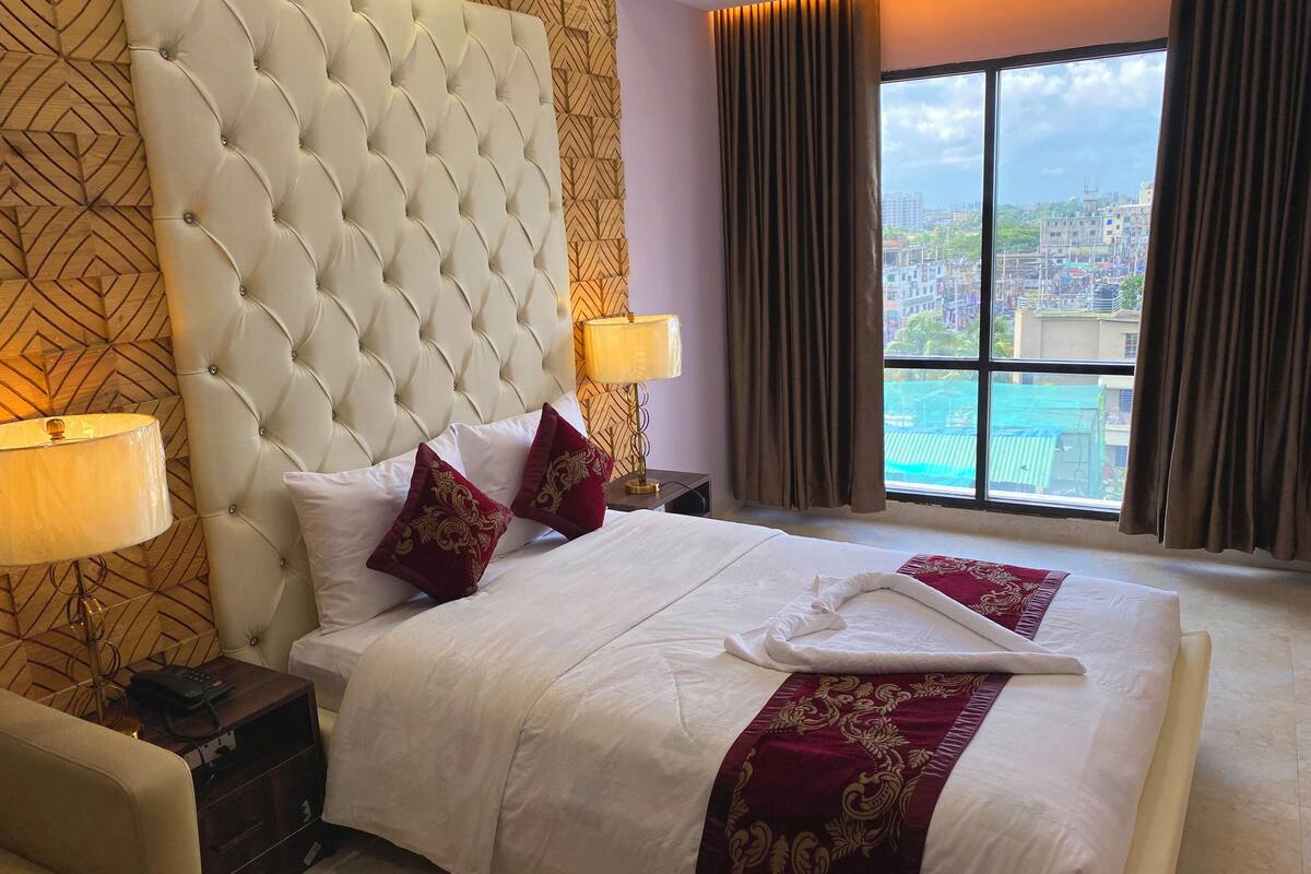 A hotel room with a tufted headboard, a large bed with white linens, two red decorative pillows, a bedside table with a lamp, and a view of a cityscape through a large window.
