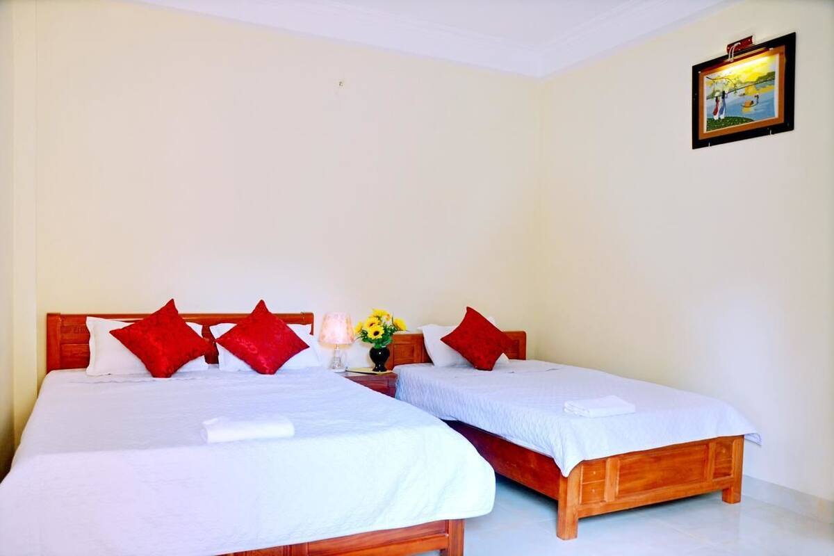 A room with two single beds, each with a wooden headboard, white bedding, and red pillows. A small table with a vase of yellow flowers is between the beds.