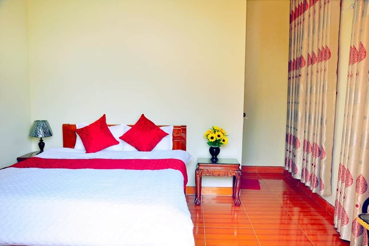 A hotel room with a bed, red pillows, a nightstand with a lamp, a vase with flowers, and curtains with a leaf pattern.