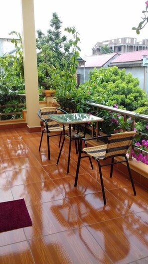 Une terrasse avec une table en verre et des chaises en osier, entourée de plantes en pot et offrant une vue sur les bâtiments.