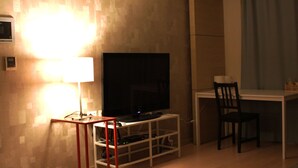 Ein Zimmer mit einem Flachbildfernseher auf einem weißen Schrank, einem kleinen Tisch mit einem Stuhl und einer Lampe auf einem roten Sockel.