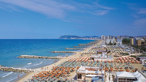 A beach with rows of umbrellas and beach chairs, a rocky breakwater, and buildings in the background. - Hotel Pensione Elena (Misano Adriatico)