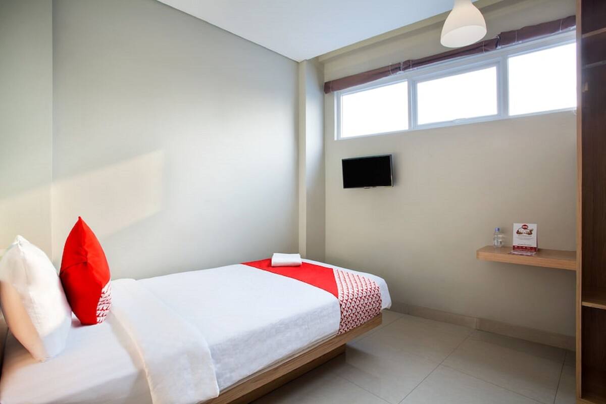 Un lit simple avec une literie blanche et des touches de rouge, un téléviseur fixé au mur et une fenêtre avec des rideaux.