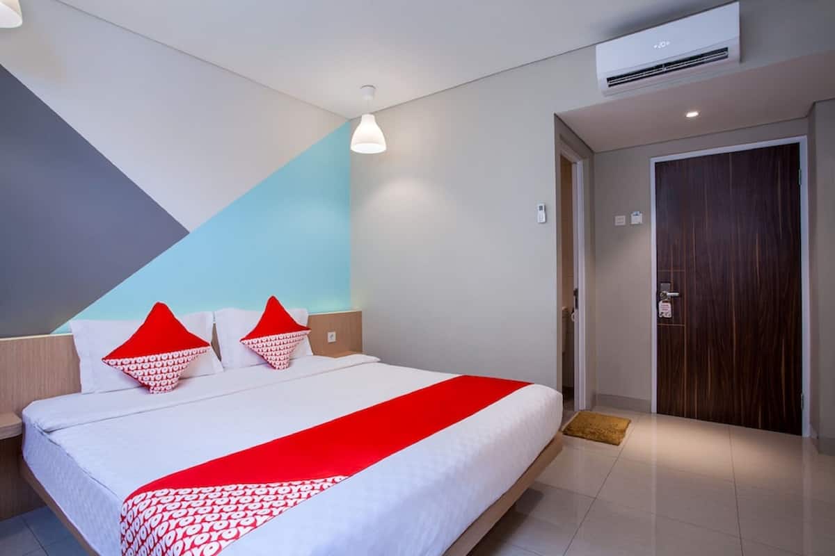Une chambre d’hôtel moderne avec un grand lit, une table de chevet, une petite table d’appoint, une couvre-lit à motifs rouges et blancs, et une porte en bois.