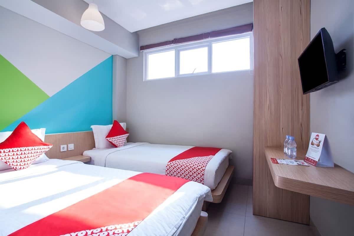 Une chambre d’hôtel avec deux lits, un téléviseur fixé au mur, une fenêtre et une étagère en bois sur laquelle se trouvent une bouteille d’eau et une brochure.