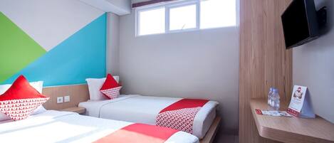 A hotel room with two beds, a TV mounted on the wall, a window, and a wooden shelf with a water bottle and a brochure.