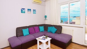 A modern living room with a sectional sofa, a small white side table, and a window with a view of the sea. - Apartments Markic (Seget)