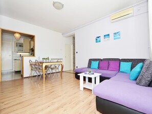 A modern living room with a purple sofa, a dining table with chairs, and a kitchen area visible through an open door.