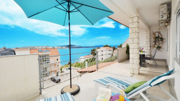 A balcony with a blue umbrella, striped cushions, and a table with drinks and a book. - Apartments Markic (Seget)