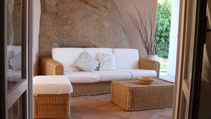 A wicker sofa with cushions, a matching ottoman, and a large rock wall in a sunlit room with a view of greenery outside. - Villa delle Rocce (Olbia)