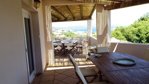 A terrace with a wooden table and chairs, a view of the sea and buildings in the distance, and a pergola overhead. - Villa delle Rocce (Olbia)