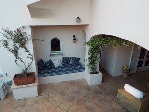 Un espacio exterior acogedor con un banco acolchado, plantas en macetas y piso de baldosas.