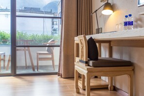 Una habitación con una mesa de madera, una silla con cojín, una ventana que ofrece vistas a zonas verdes y edificios, y una lámpara sobre la mesa.