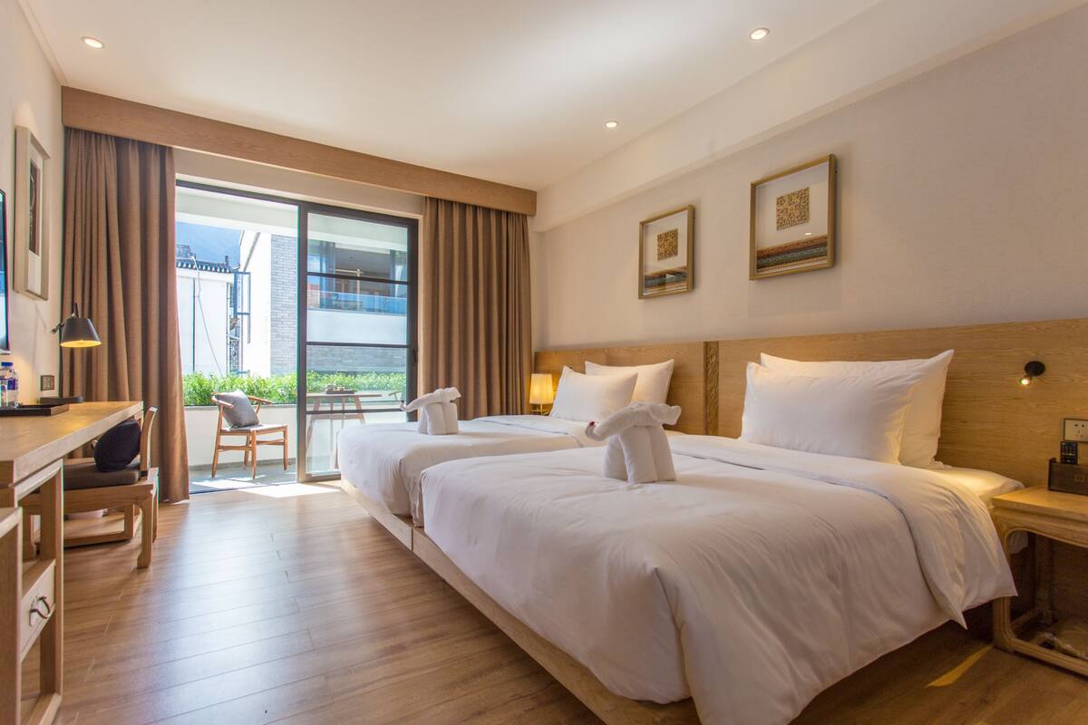 Habitación de hotel con dos camas, un escritorio, una silla y un ventanal con cortinas.