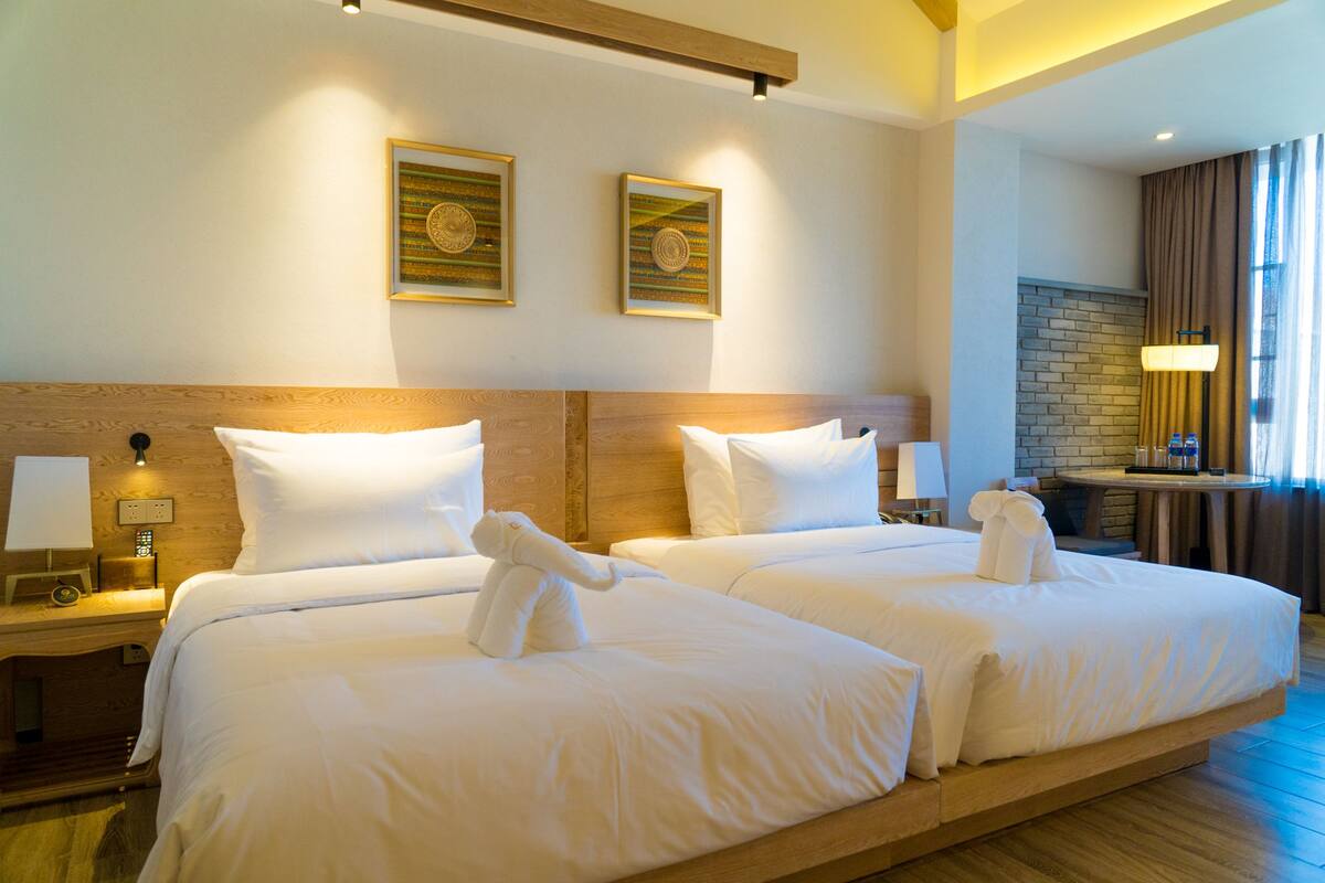 Habitación de hotel con dos camas, cabecero de madera, pared de ladrillo y ventana con cortinas.