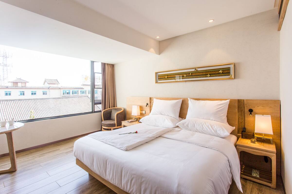 Habitación de hotel con una cama grande, una mesita de noche con lámpara, una silla y vistas a edificios a través de la ventana.