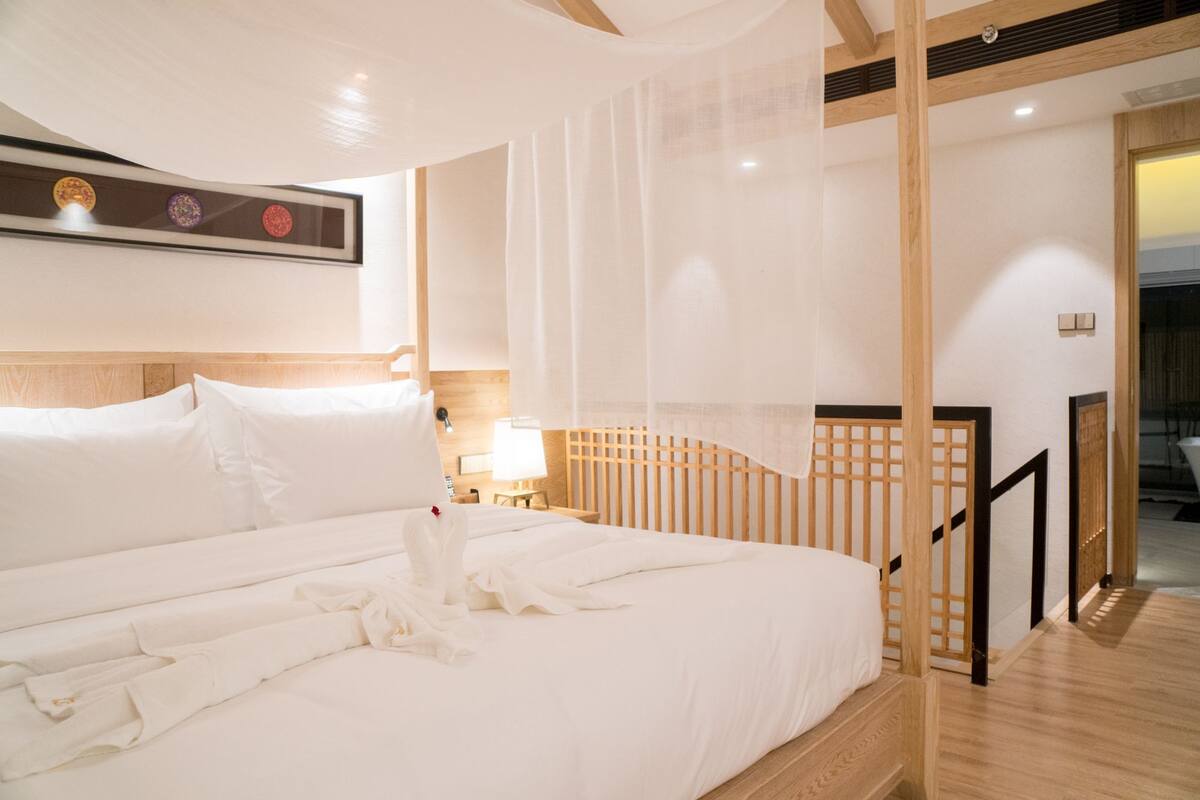 Una habitación de hotel con una cama grande, un cabecero de madera, una mesita de noche con una lámpara y vistas al exterior a través de una ventana.