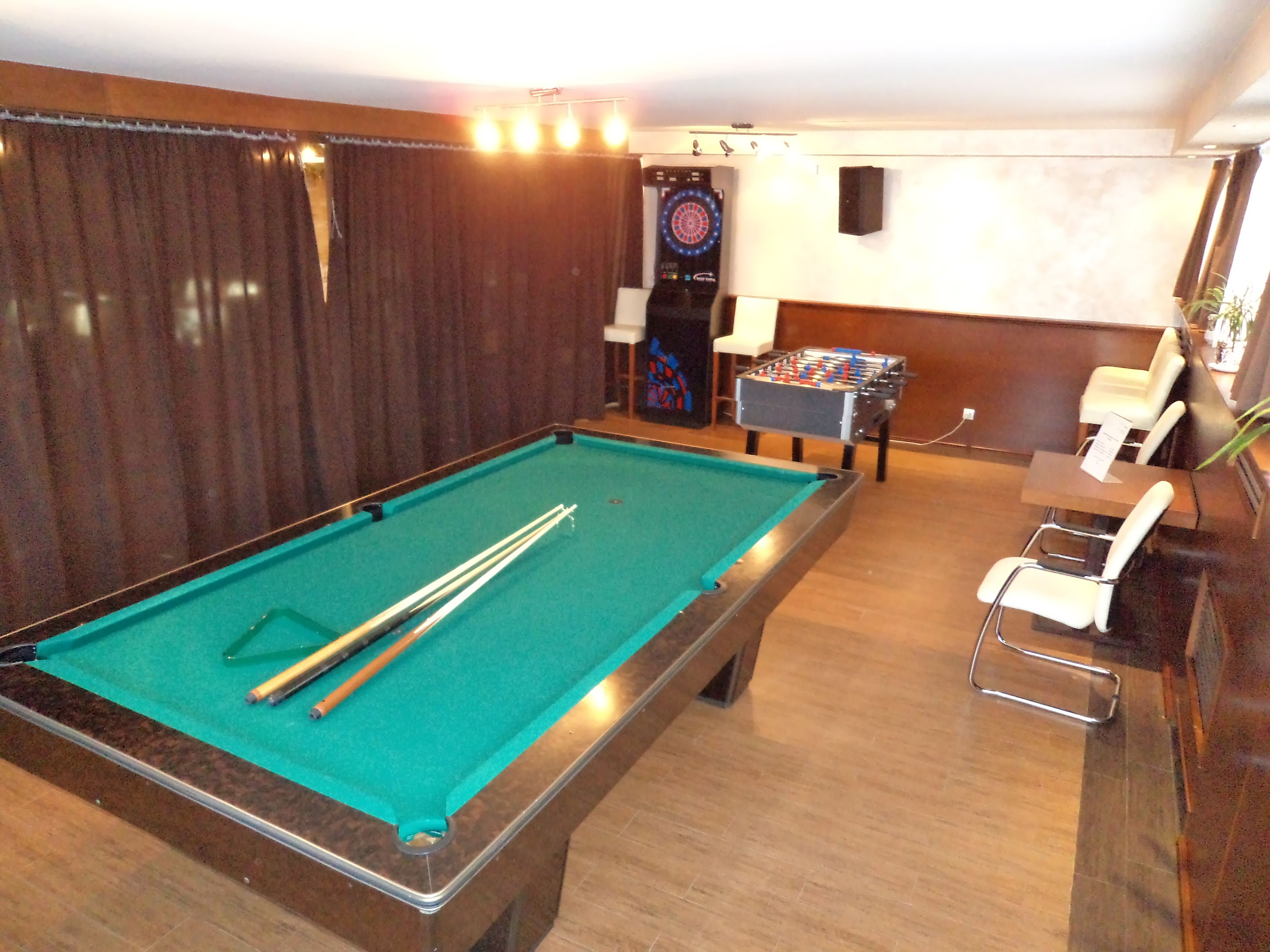 A pool table with pool cues, a foosball table, and a dartboard in a room with wooden flooring and a brown curtain.