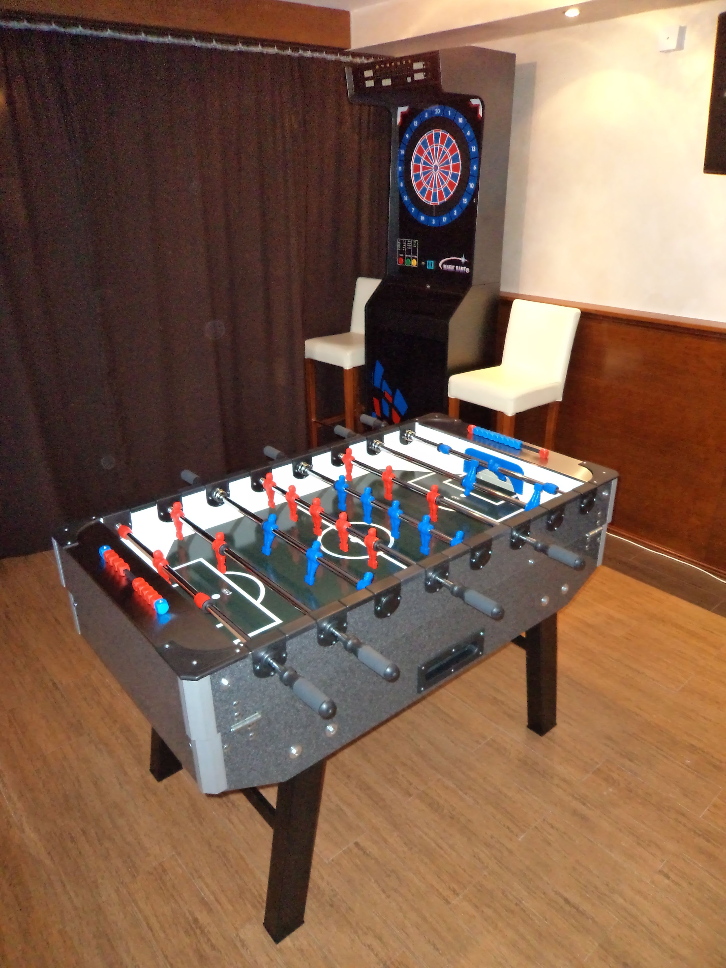 Une table de baby-foot dans une pièce où se trouvent également un jeu de fléchettes et une chaise.