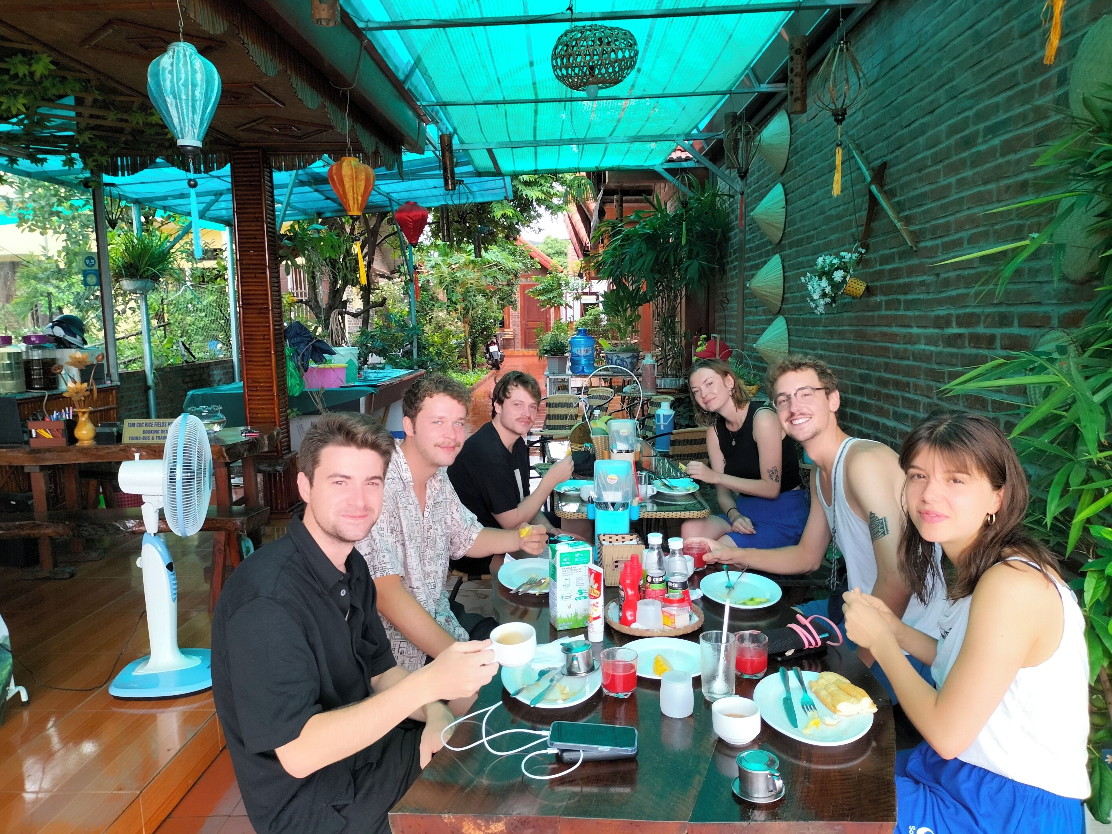 A group of people dining at an outdoor restaurant with tables, chairs, and various food and drink items.