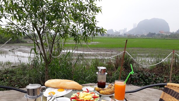 A table set with breakfast items including eggs, toast, fruit, and beverages, overlooking a rural landscape with fields and a distant mountain. - Tam Coc Rice Fields Homestay (Nam Hoa Lu)