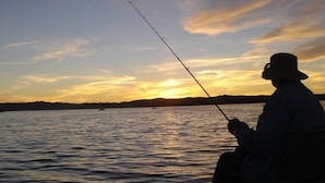 Pessoa pescando ao pôr do sol, com vista clara da água e da costa distante.