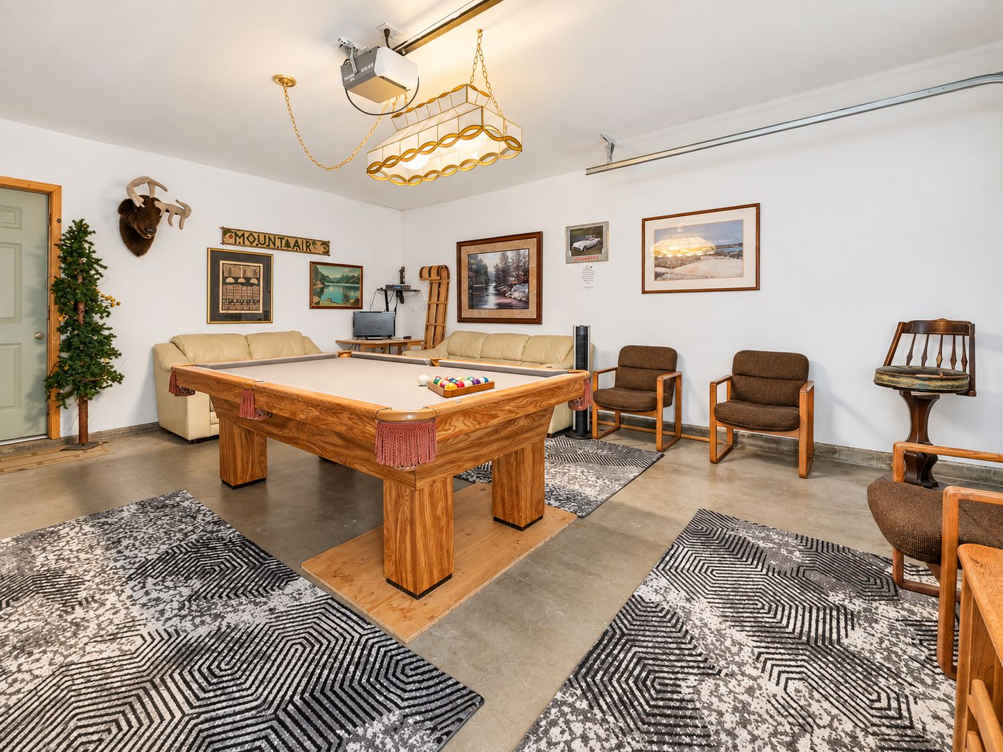 Una habitación con mesa de billar, sofá y una silla. Se observa un trofeo de venado montado en la pared y un cuadro enmarcado.