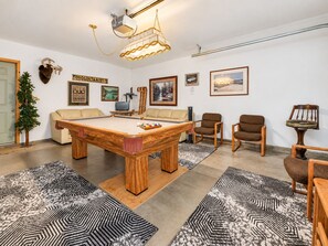 Une pièce comprenant une table de billard, un canapé et un fauteuil. Un trophée de tête de cerf est accroché au mur, ainsi qu’un tableau encadré.