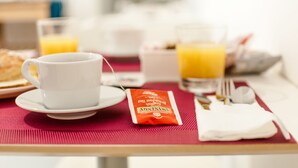 A breakfast setting with a white cup, a glass of orange juice, a packet of instant coffee, and a plate of food on a red tablecloth. - Speciale Guesthouse (Rome)