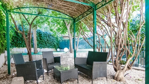 A covered outdoor seating area with wicker furniture and a glass table, surrounded by trees and greenery. - Speciale Guesthouse (Rome)
