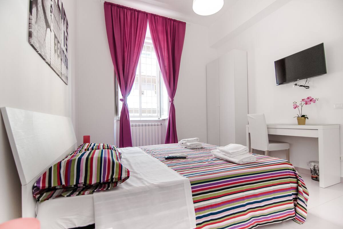 A hotel room with a bed, a desk, a TV, and a window with curtains.