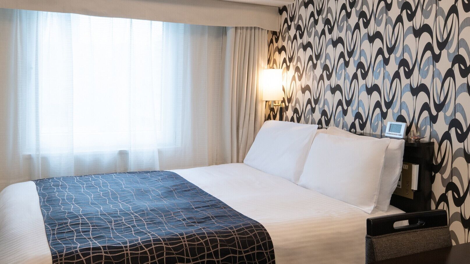 A hotel room with a bed, a bedside table with a lamp, and a wall with a patterned wallpaper.