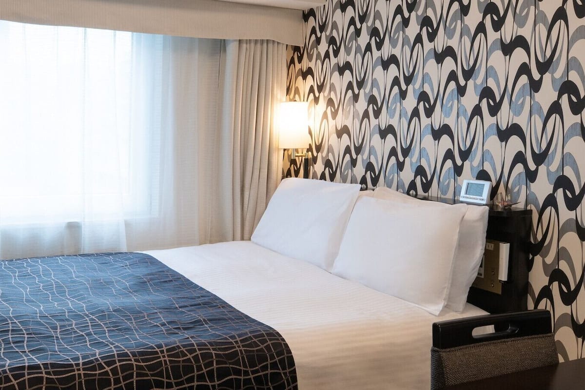 A hotel room with a bed, a bedside table with a lamp, and a wall with a patterned wallpaper.