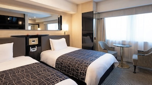 Quarto de hotel com duas camas, uma escrivaninha, uma cadeira, uma mesinha, um espelho e uma janela com cortinas.