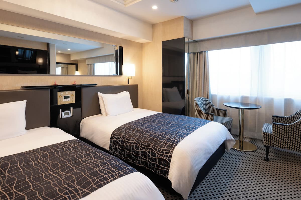 Ein Hotelzimmer mit zwei Betten, einem Schreibtisch, einem Stuhl, einem kleinen Tisch, einem Spiegel und einem Fenster mit Vorhängen.