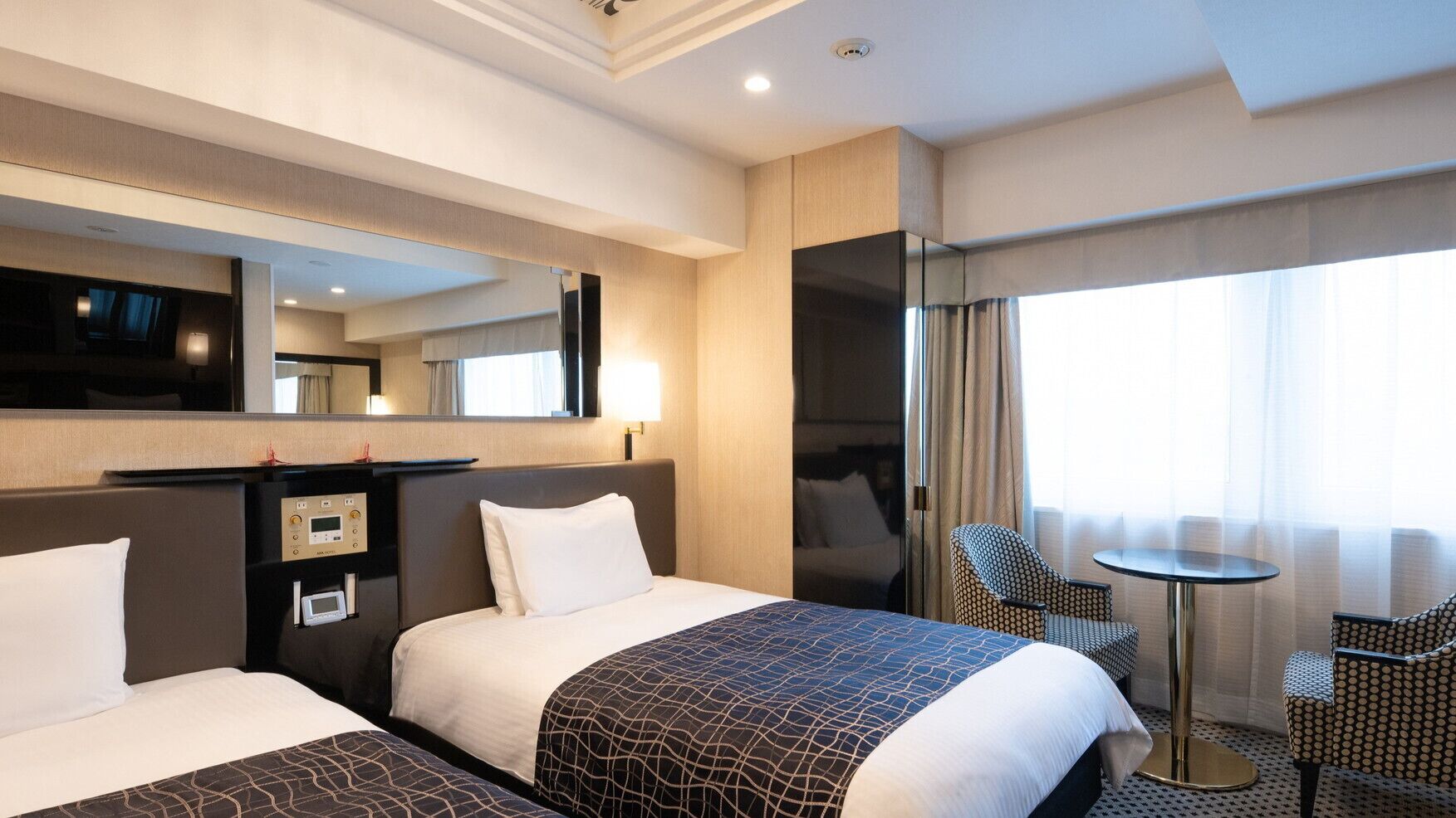 A hotel room with two beds, a desk with a control panel, a chair, a small table, and a large mirror.