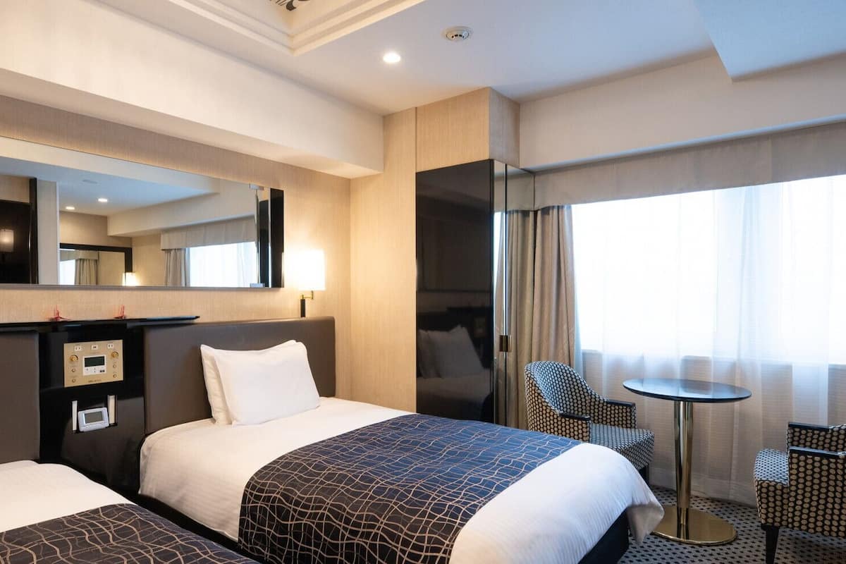 A hotel room with two beds, a desk with a control panel, a chair, a small table, and a large mirror.