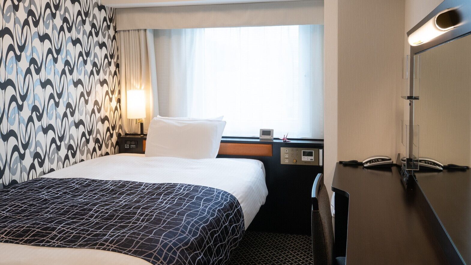 A hotel room with a bed, a desk with a phone, a control panel, and a window with curtains.