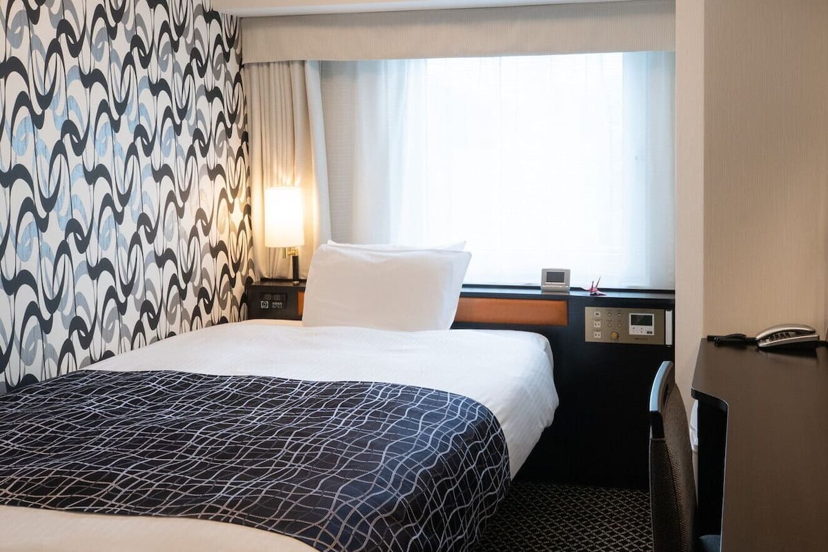 A hotel room with a bed, a desk with a phone, a control panel, and a window with curtains.