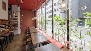 Um restaurante com longas mesas e bancos de madeira, parede de tijolos e vista para a rua através de grandes janelas.