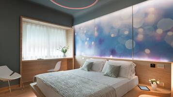 A modern bedroom with a large bed, a bedside table with a tablet, a chair, and a window with sheer curtains.
