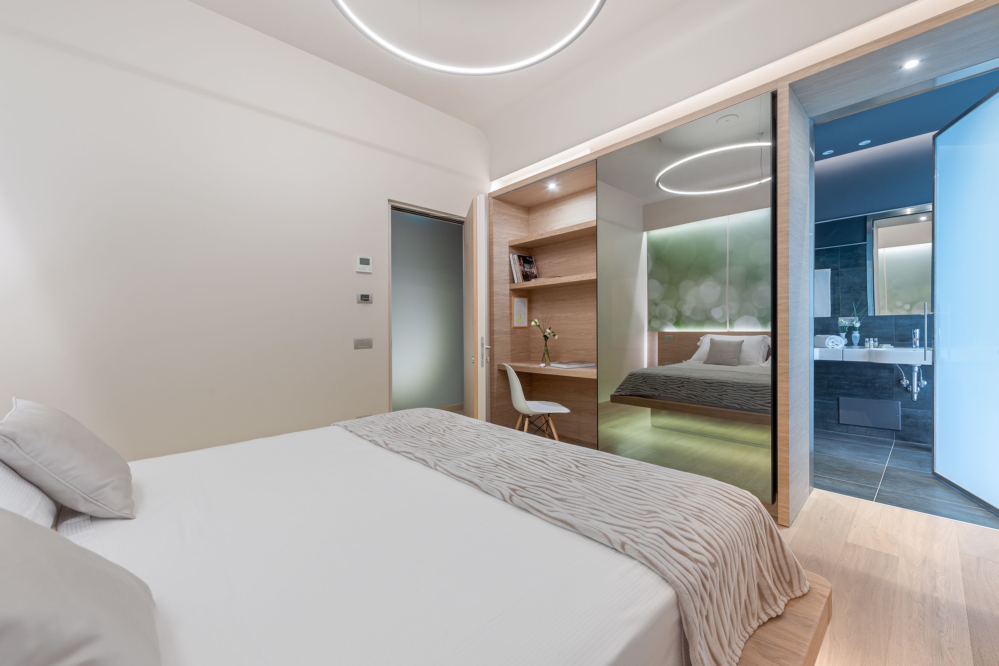 Camera da letto moderna con un letto grande, una scrivania e un bagno visibile attraverso una porta in vetro.