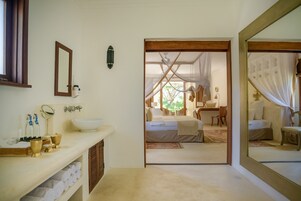 A bathroom with a sink, mirror, and toiletries. There is a doorway leading to a bedroom with a bed and a window.