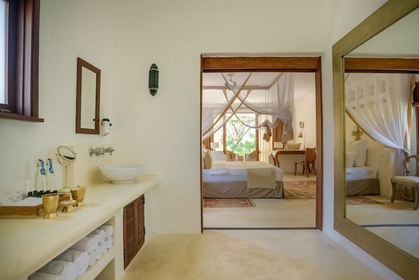 A bathroom with a sink, mirror, and toiletries. There is a doorway leading to a bedroom with a bed and a window. - Swahili Beach Resort (Diani Beach)