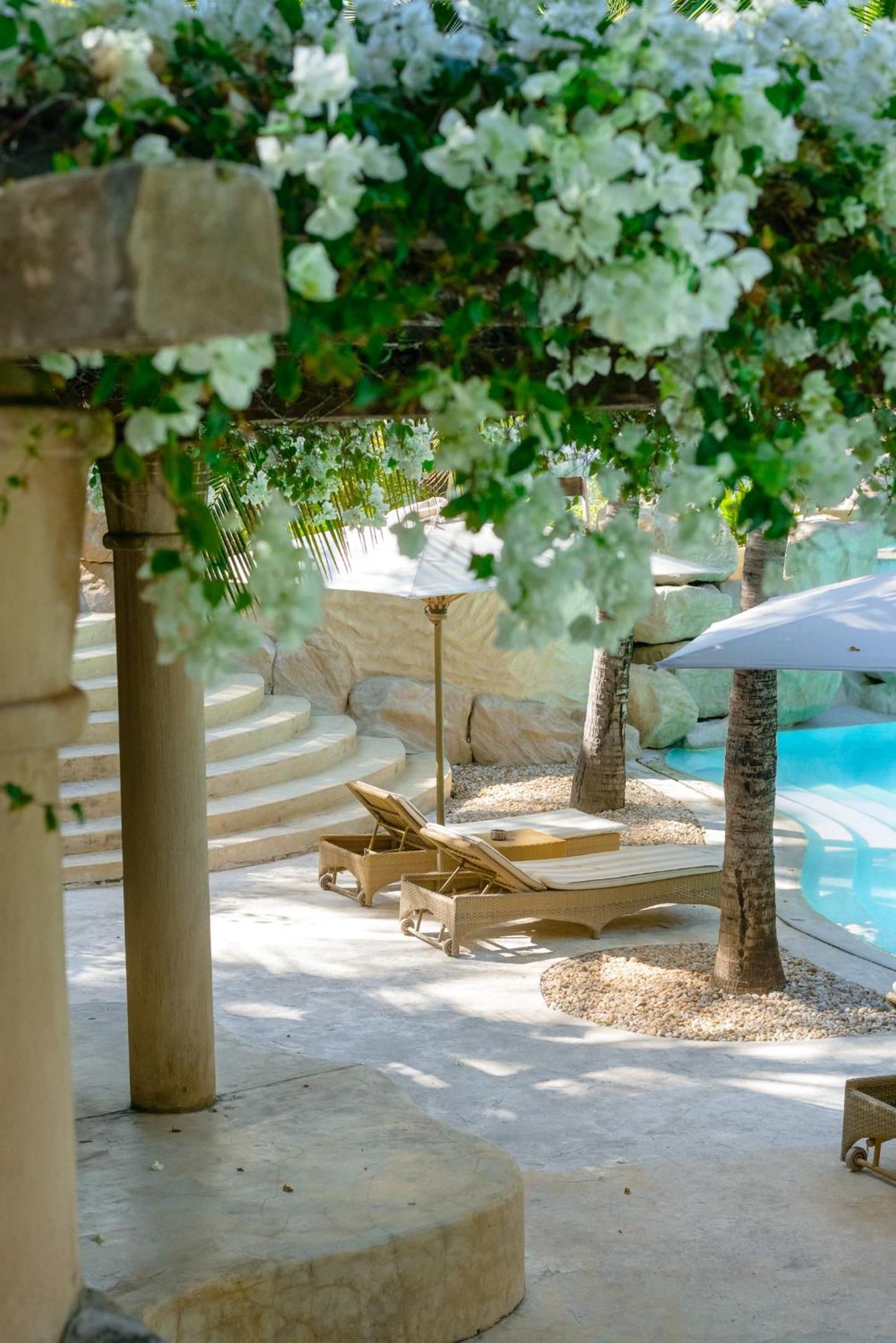 A poolside area with lounge chairs, a stone wall, and a pergola with white flowers.