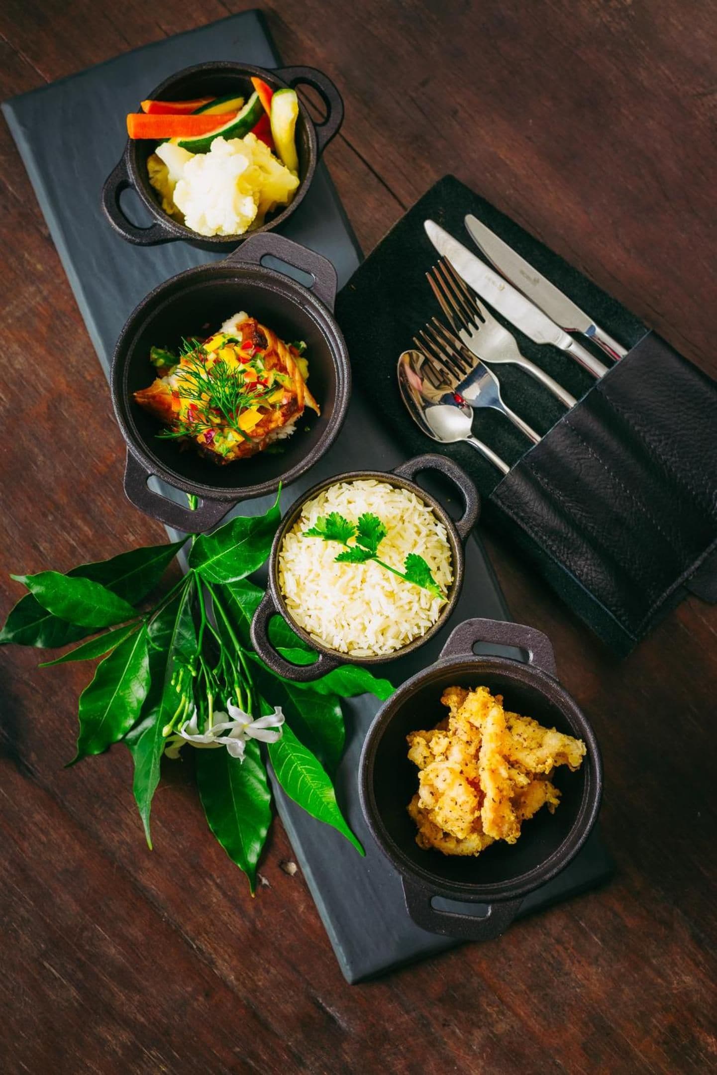 Une table avec quatre récipients noirs contenant différents plats, une serviette noire avec des couverts, et une plante aux feuilles vertes.