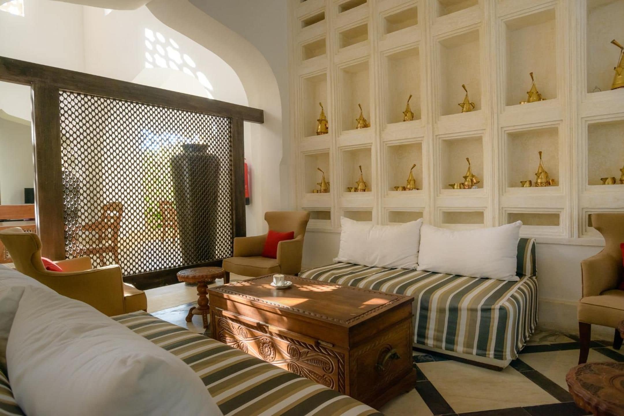 A room with a sofa, armchairs, a wooden chest, and a wall with shelves holding decorative items.